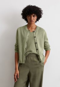 Hellgrüne Strickjacke mit rundem Halsausschnitt, vorderem Knopfverschluss und langen Ärmeln, über einem passenden strukturierten Oberteil und olivgrünen Hosen getragen.