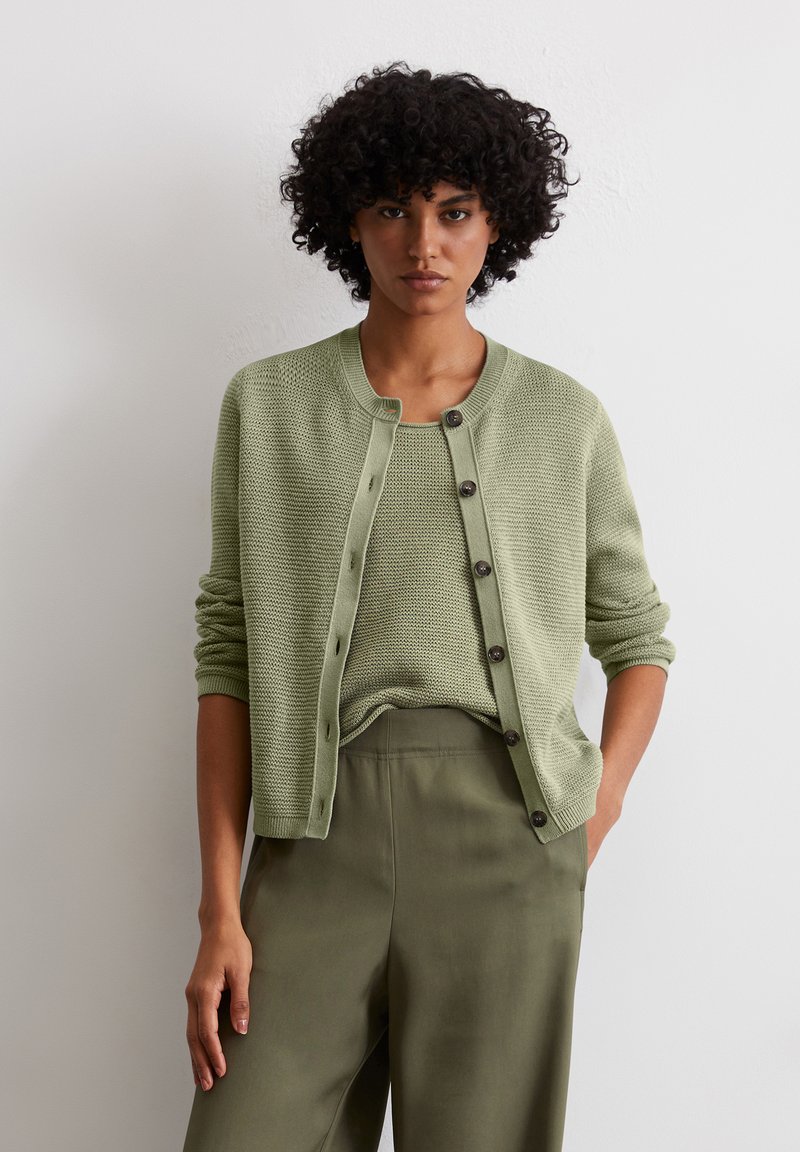 Hellgrüne Strickjacke mit rundem Halsausschnitt, vorderem Knopfverschluss und langen Ärmeln, über einem passenden strukturierten Oberteil und olivgrünen Hosen getragen.