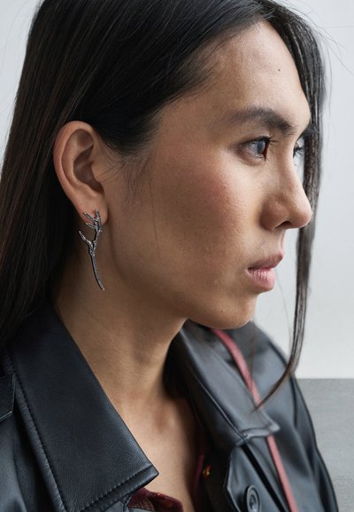 Ασημένιο σκουλαρίκι σε σχήμα κλαδιού, με λεπτομερείς, υφασμένους τόνους· φορεμένο από μοντέλο με σκούρα μαλλιά, πάνω σε ουδέτερο φόντο.