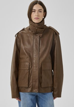 Frau trägt eine braune Lederjacke mit hohem Kragen und großen Vordertaschen, kombiniert mit blauen Jeans, stehend vor einfachem Hintergrund.