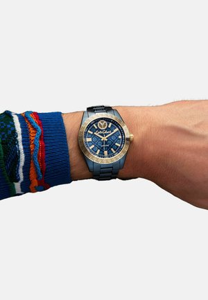 Blaues Zifferblatt-Armbanduhr mit goldenen Akzenten, gemusterter Lünette und schwarzem Armband. Am Handgelenk getragen mit einem Pullover, der farbenfrohe Muster aufweist.
