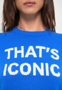 Blå strikkegenser med hvit tekst som sier "THAT'S ICONIC." Den har rund hals og ribbet tekstur.