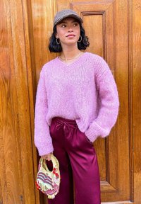 Pull en laine rose et doux avec un col en V, accompagné de pantalons en satin bordeaux. Tenant un petit sac à main coloré et brodé. Fond en bois.