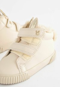 Baskets montantes en simili cuir beige avec deux bandes Velcro, doublure duveteuse et accent métallique en forme de lapin sur la sangle. Semelle en caoutchouc texturée.
