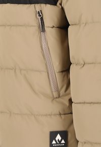 Beige donsjacket met een zwart bovenstuk. Bevat een verticale ritszak en een zwart logo patch met een witte bergafbeelding.