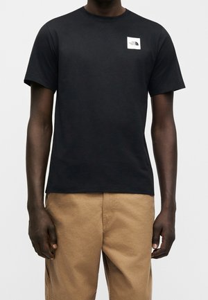 Homme portant un T-shirt noir à manches courtes avec un petit logo blanc sur la poitrine gauche et un pantalon marron clair sur un fond uni.
