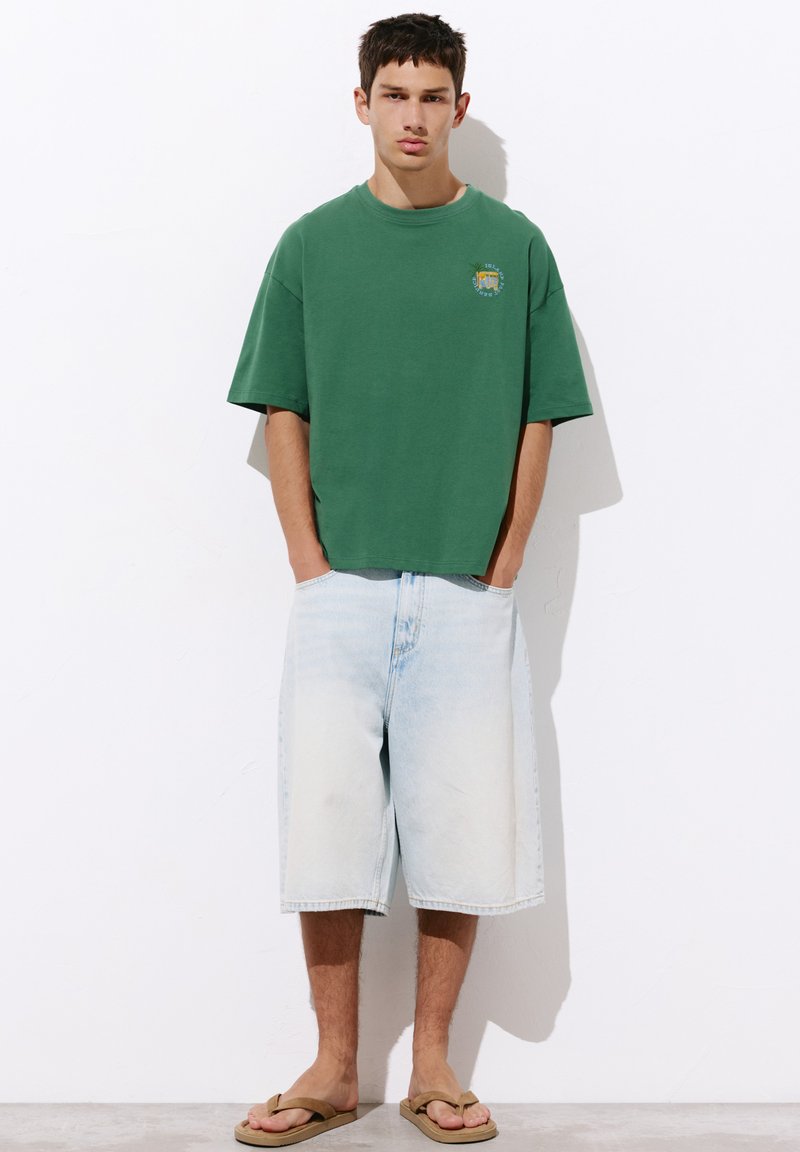 Jeune homme debout contre un mur blanc, portant un T-shirt vert ample, un long short en jean clair et des sandales brunes, les mains dans les poches.
