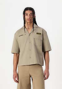 Camicia khaki a maniche corte con chiusura a bottoni, tasca sul petto e testo ricamato. Tessuto morbido e design rilassato.