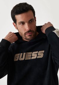 Čierna mikina s kapucňou a zlatým logom "GUESS", s viazaním na kapucni, rebrovanými manžetami a kontrastným detailom na rukávoch. Mäkká textúra.