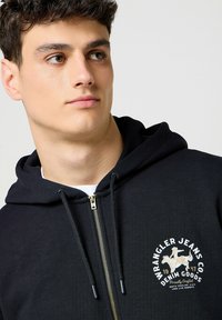 Black zip-up hoodie made of soft fabric, featuring a logo patch in white and gold at the chest, with drawstrings and a kangaroo pocket.