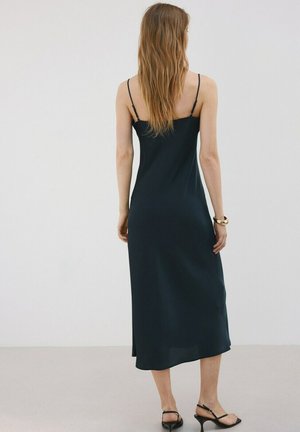 Mujer de cabello largo que lleva un vestido midi negro sin mangas con tirantes delgados y tacones abiertos negros, de pie frente a un fondo liso.