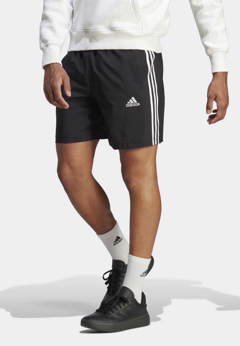 Schwarze Sportshorts mit einem weißen Dreistreifen-Design, elastischem Bund und leichtem Stoff. Schwarze Sneakers und weiße Socken mit sichtbarem Logo.