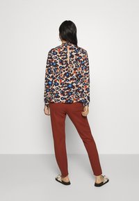 Dlouhý rukáv blouse v béžové barvě s barevným abstraktním potiskem; má výstřih ve tvaru klíčové dírky na zádech a elastické manžety, skombinováno s kalhotami v barvě rez.