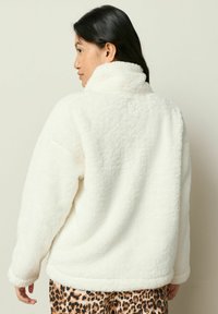 Hvid fleecejakke med høj krave og blød tekstur, der har en afslappet pasform og ribbet kant. Sammen med leopardmønstrede bukser.