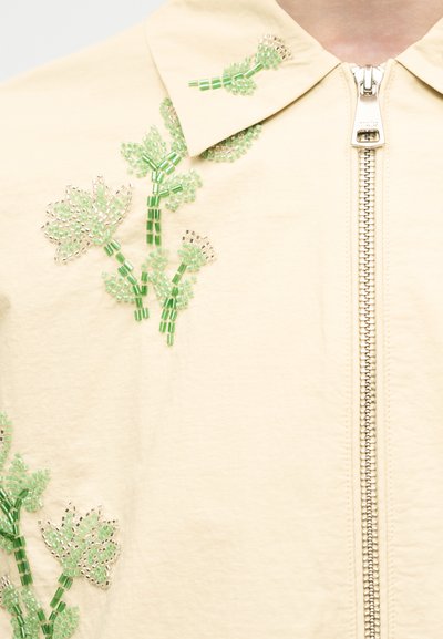 Ύφασμα εκρού με πράσινη και ασημένια κεντητή κλωστή σε σχέδιο λουλουδιών κοντά στον γιακά και το μπροστινό φερμουάρ ενός ενδύματος.