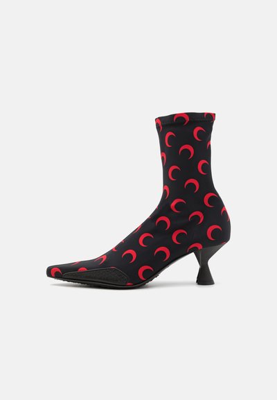 Botín negro de tobillo en tela elástica, con patrones de lunas crecientes rojas y punta afilada. Un pequeño tacón negro angular completa el diseño.