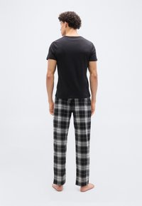 Άνδρας που στέκεται με γυμνά πόδια γυρισμένος προς τα πίσω, φορώντας μαύρο κοντομάνικο πουκάμισο και μαύρο-λευκό καρό παντελόνι, με απλό φόντο.