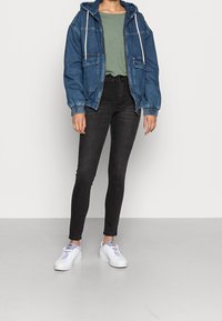 Jeansjacka med huva, två framfickor och elastiska ärmar, kombinerad med en grön t-shirt och svarta skinny jeans samt vita sneakers.