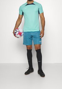 Camiseta deportiva Nike de color azul claro con acentos en azul oscuro, combinada con pantalones cortos turquesa y calcetines negros. Sosteniendo un balón de fútbol con diseños coloridos.