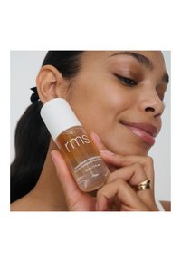 Klare Glasflasche mit weißem Deckel, gefüllt mit Flüssigkeitsserum. Auf dem Etikett steht "rms Super Hydrating Serum." Das Modell hält die Flasche nah am Gesicht.
