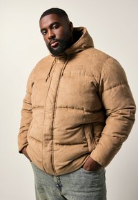 Hombre con barba que lleva una chaqueta acolchada beige con capucha y vaqueros azules, de pie con las manos en los bolsillos, fondo neutro.
