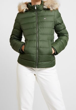 Personne portant une veste matelassée verte avec une bordure en fourrure sur la capuche et un pantalon blanc, les mains dans les poches avant, debout devant un fond uni.