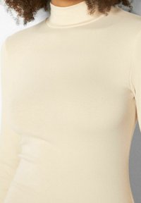 Beige pitkähihaisen poolopaidan toppi, valmistettu sileästä, joustavasta materiaalista. Varustettu vartalonmyötäisellä leikkauksella, korkealla kaula-aukolla ja saumattomalla rakenteella.