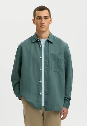 Młody mężczyzna w luźnej turkusowej koszuli na guziki, założonej na białą koszulkę, oraz beżowych spodniach, stojący na tle jasnego, jednolitego tła.