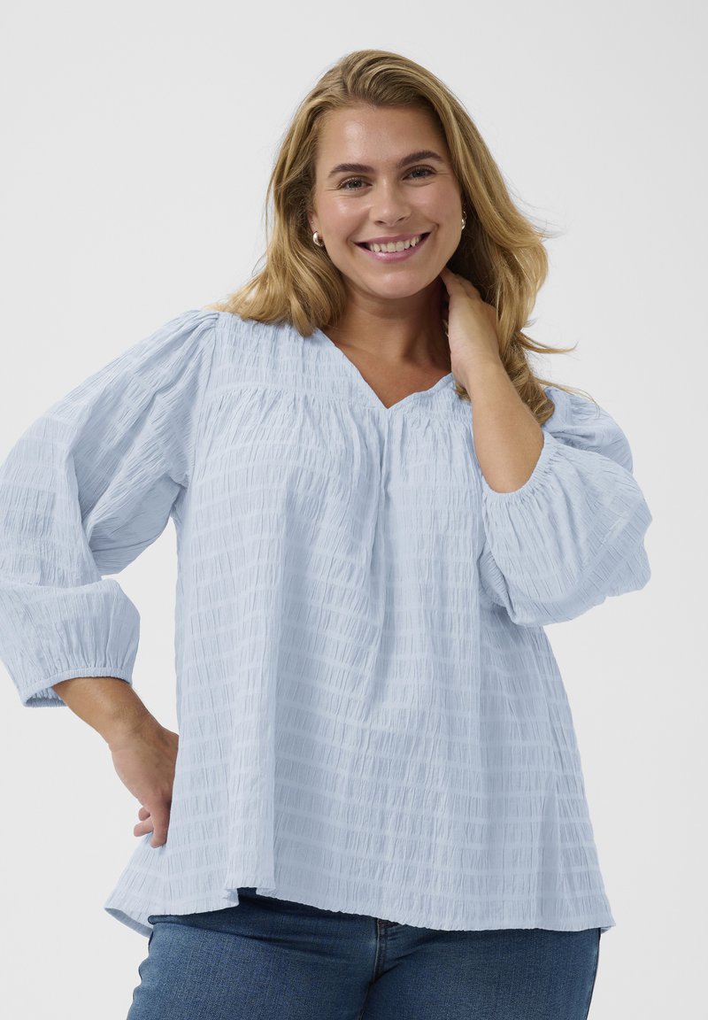 Femme souriante aux longs cheveux blonds portant un chemisier bleu clair texturé et un jean bleu, posant avec une main sur le cou sur fond blanc.
