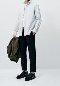 Camicia azzurro chiaro con bottoni, maglietta bianca a girocollo, pantaloni scuri, scarpe nere stringate, che tiene in mano una giacca verde oliva.