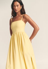 Keltainen midimekko, jossa neliskanttinen pääntie, ohuet olkaimet ja sivuhiuskat; valmistettu kevyestä, sileän tuntuista kankaasta.