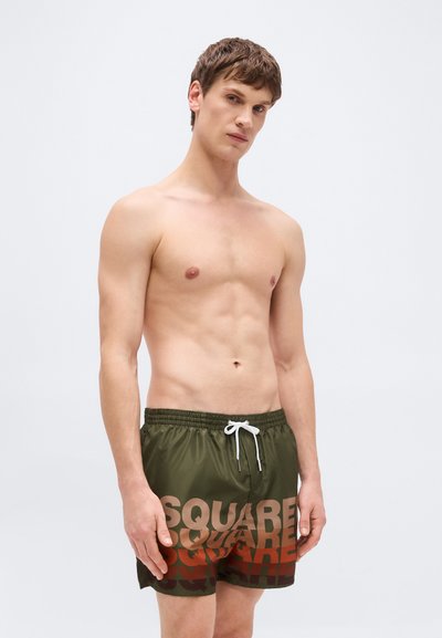 Νέος άνδρας με κοντά καστανά μαλλιά στέκεται γυμνόστηθος φορώντας χακί σορτς κολύμβησης με το κείμενο "SQUARE" σε αποχρώσεις καφέ-κόκκινου.