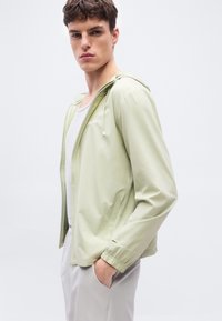 Νέος άνδρας που φοράει φούτερ με φερμουάρ ανοιχτόχρωμου πράσινου πάνω από λευκή φανέλα χωρίς μανίκια και ανοιχτό γκρι παντελόνι, πόζαροντας με το ένα χέρι στην τσέπη μπροστά σε απλό φόντο.