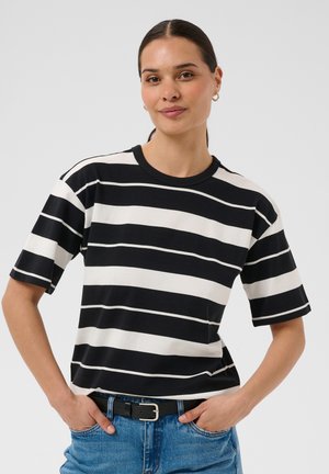 Femme aux cheveux foncés attachés porte un t-shirt à rayures horizontales noires et blanches à manches courtes, un jean bleu et une ceinture noire, debout, les mains dans les poches.