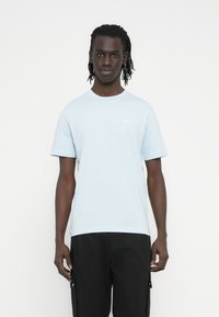 Ανοιχτό μπλε βαμβακερό μπλουζάκι με στρογγυλή λαιμόκοψη, κοντά μανίκια και μικρό λευκό λογότυπο στο στήθος. Φοράει μαύρο παντελόνι.