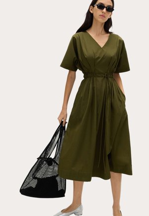 Kvinde iført en olivengrøn midi-kjole med V-hals og taljeret talje, bærende en stor sort net-tote taske, iført sorte solbriller og sølvfarvede flade sko.
