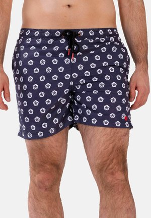Mann trägt marineblaue Badeshorts mit weißem Blumenmuster und schwarzem Kordelzug, ab Taille bis Mitte Oberschenkel vor weißem Hintergrund gezeigt.