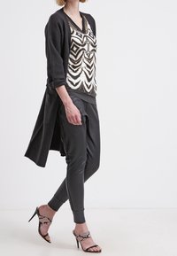 Černý dlouhý kardigan přes vzorovaný bílý a černý top, spárovaný s černými skinny kalhotami a vysokými sandály s hadím vzorem na popruhu.