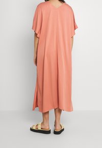 Langes, locker sitzendes Kleid in Koralle mit V-Ausschnitt und kurzen Ärmeln. Der Stoff hat eine glatte Textur, ideal für warmes Wetter. Kombiniert mit klobigen Sandalen.