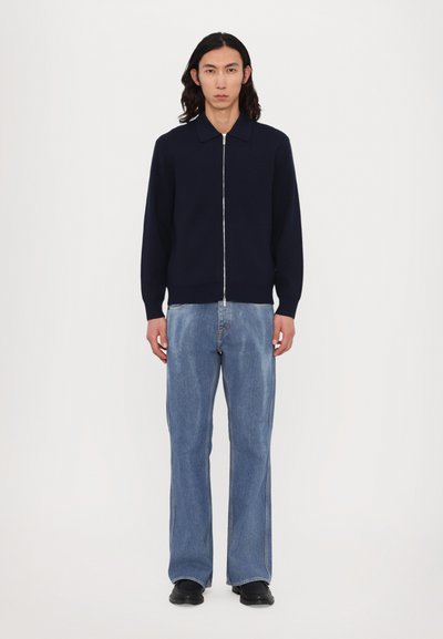 Cardigan azul-marinho com fecho, em malha suave, combinado com jeans claros de corte flare e sapatos pretos. Design simples, com poucos detalhes e cores neutras.