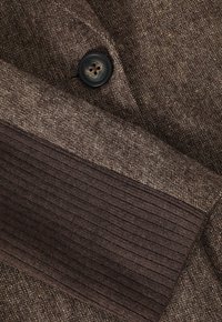 Ruskea tekstuuri kangas pystysuuntaisella ribbineuleosalla, jossa on neljällä reiällä varustettu musta nappi. Kokonaisvärimaailma on tummanruskea.