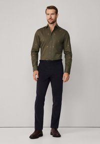 Man die tegen een effen achtergrond staat, gekleed in een olijfgroen versierde shirt, zwarte broek en donkerbruine schoenen.