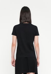 Persona con capelli scuri e lisci che indossa una semplice maglietta nera a maniche corte e una gonna nera, vista di spalle su uno sfondo bianco.