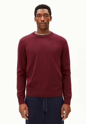 Mann trägt einen eng anliegenden bordeauxroten Langarmpullover und dunkelblaue Hose, steht vor einem schlichten hellen Hintergrund.