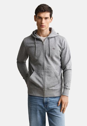 Młody mężczyzna ubrany w jasnoszarą bluzę z kapturem na zamek błyskawiczny z przednimi kieszeniami oraz jasnoniebieskie dżinsy, stojący na jednolitym białym tle.