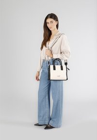 Czarna i biała torebka z górnymi uchwytami oraz odpinanym paskiem na ramię. Modelka ubrana w jasną koszulę z guzikami i spodnie z szerokimi nogawkami.