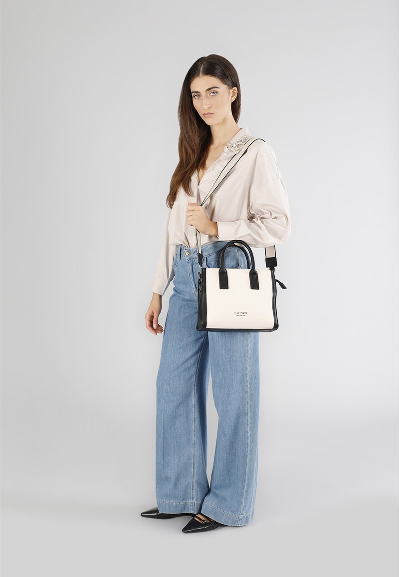Czarna i biała torebka z górnymi uchwytami oraz odpinanym paskiem na ramię. Modelka ubrana w jasną koszulę z guzikami i spodnie z szerokimi nogawkami.