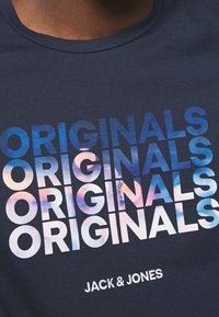 Námořnická modrá košile s opakujícím se textem "ORIGINALS" v modré, růžové a bílé gradaci a logem "JACK & JONES" níže.