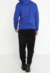Sudadera azul con capucha de tejido texturizado, combinada con pantalones de chándal negros con rayas laterales y zapatos negros. Vista desde atrás.
