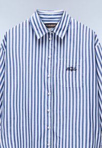 Camicia a righe blu e bianca in cotone. Presenta un colletto classico, taschino frontale e logo ricamato sulla tasca.